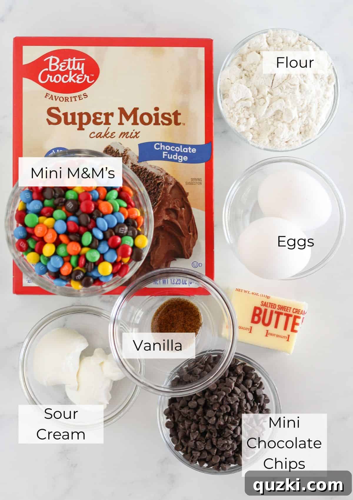 Ultimate Chocolate M and Ms Cookie Treat 3 A collection of ingredients laid out on a surface, including a box of chocolate cake mix, flour, eggs, butter, sour cream, vanilla, mini chocolate chips, and mini M&M candies, all ready to make cookies.