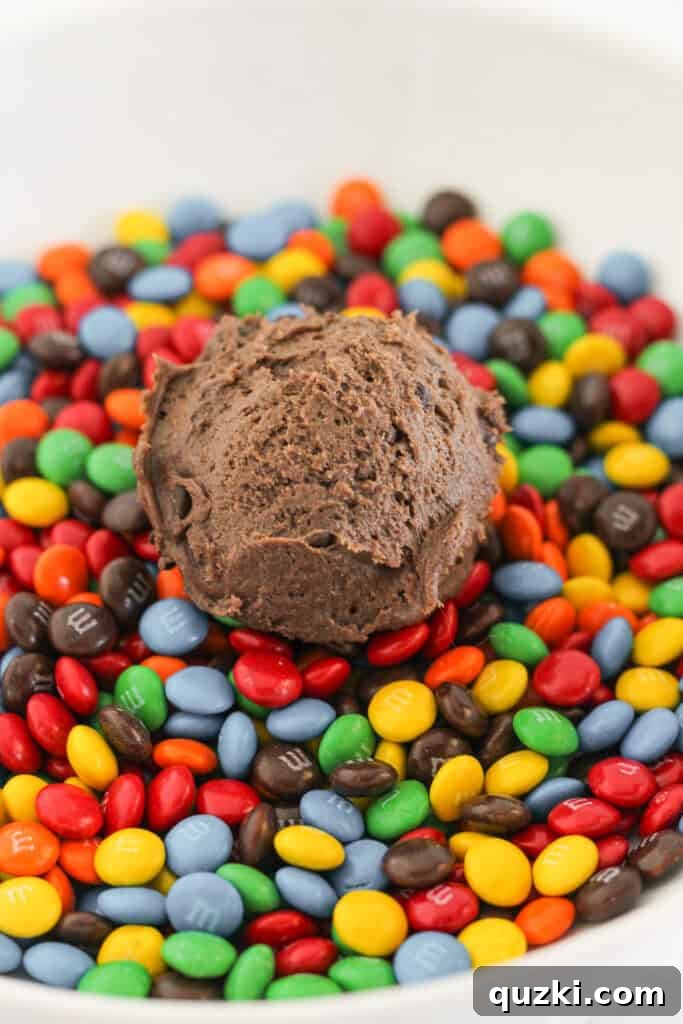 Ultimate Chocolate M and Ms Cookie Treat 5 A portion of chocolate cookie dough, expertly scooped into a ball, poised just above a pile of colorful mini M&M candies, ready to be rolled.