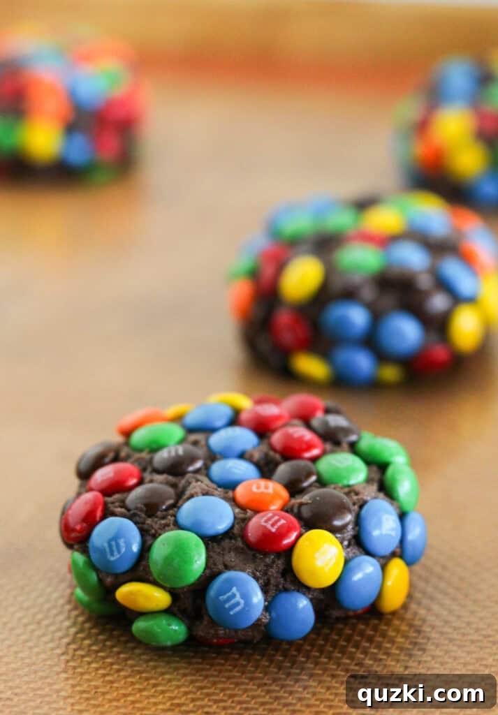 Ultimate Chocolate M and Ms Cookie Treat 6 Rows of chocolate cake mix cookie dough balls, generously coated in mini M&M candies, neatly arranged on a baking sheet lined with parchment paper, ready for the oven.
