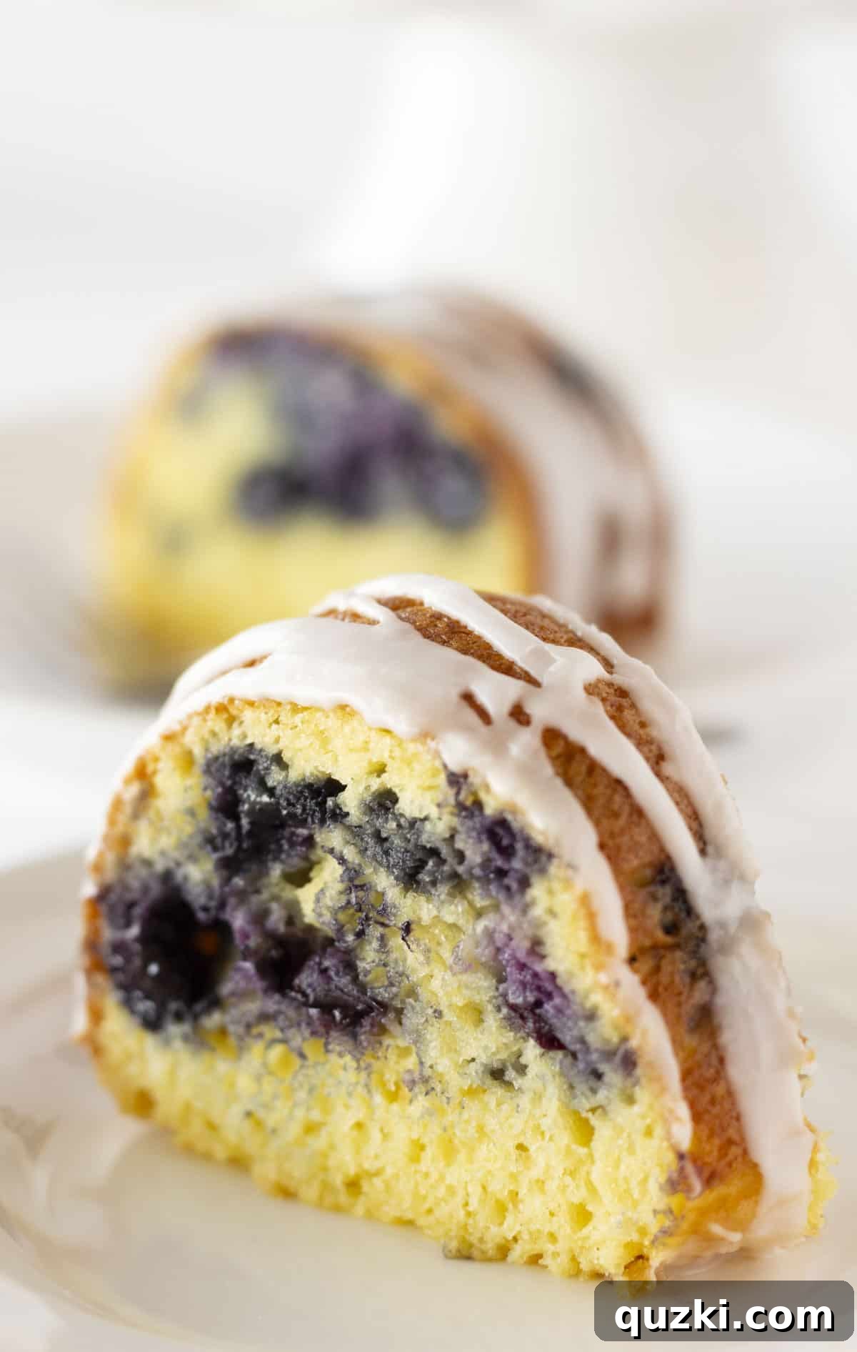 Two generous slices of blueberry bundt cake on a white plate, showcasing the fluffy texture and visible blueberries, drizzled with glaze and ready to be enjoyed.