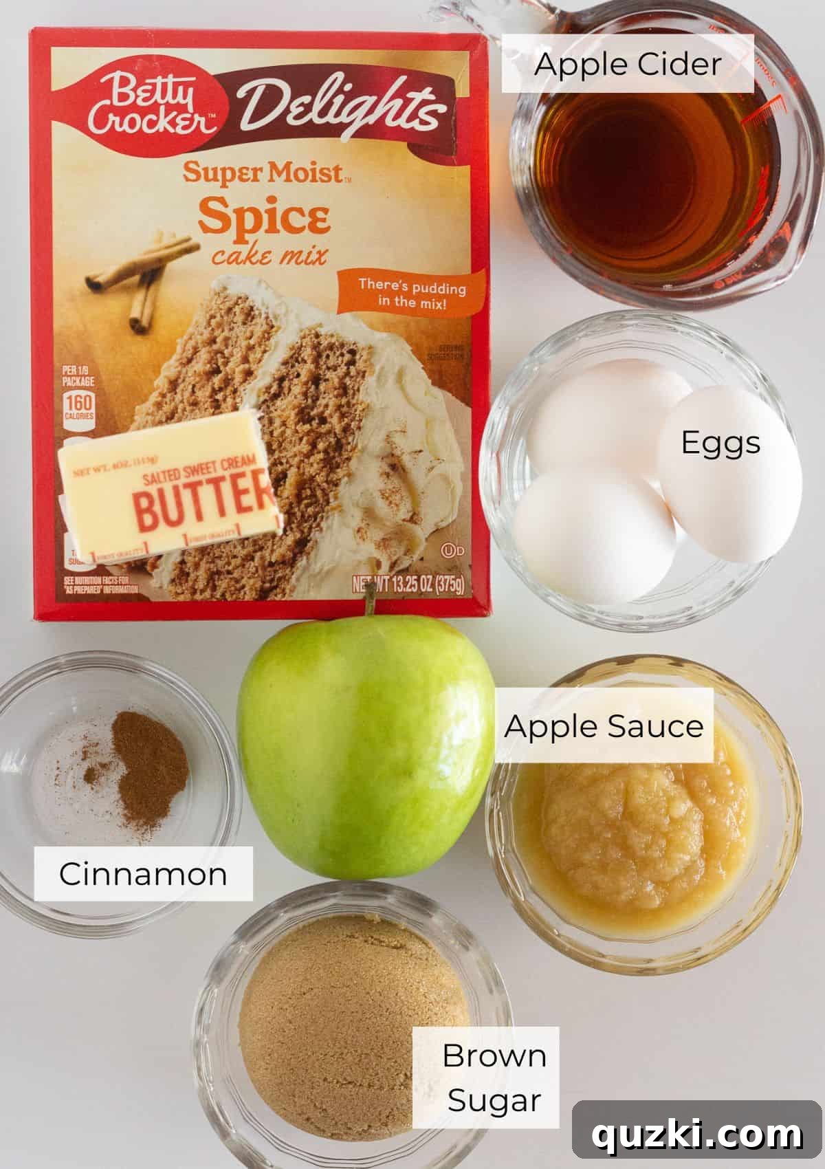 No-Fuss Caramel Apple Upside Down Cake 3 A collection of ingredients laid out on a table, including apples, butter, brown sugar, cinnamon, a box of spice cake mix, apple cider, and applesauce, ready for making an apple upside down cake.