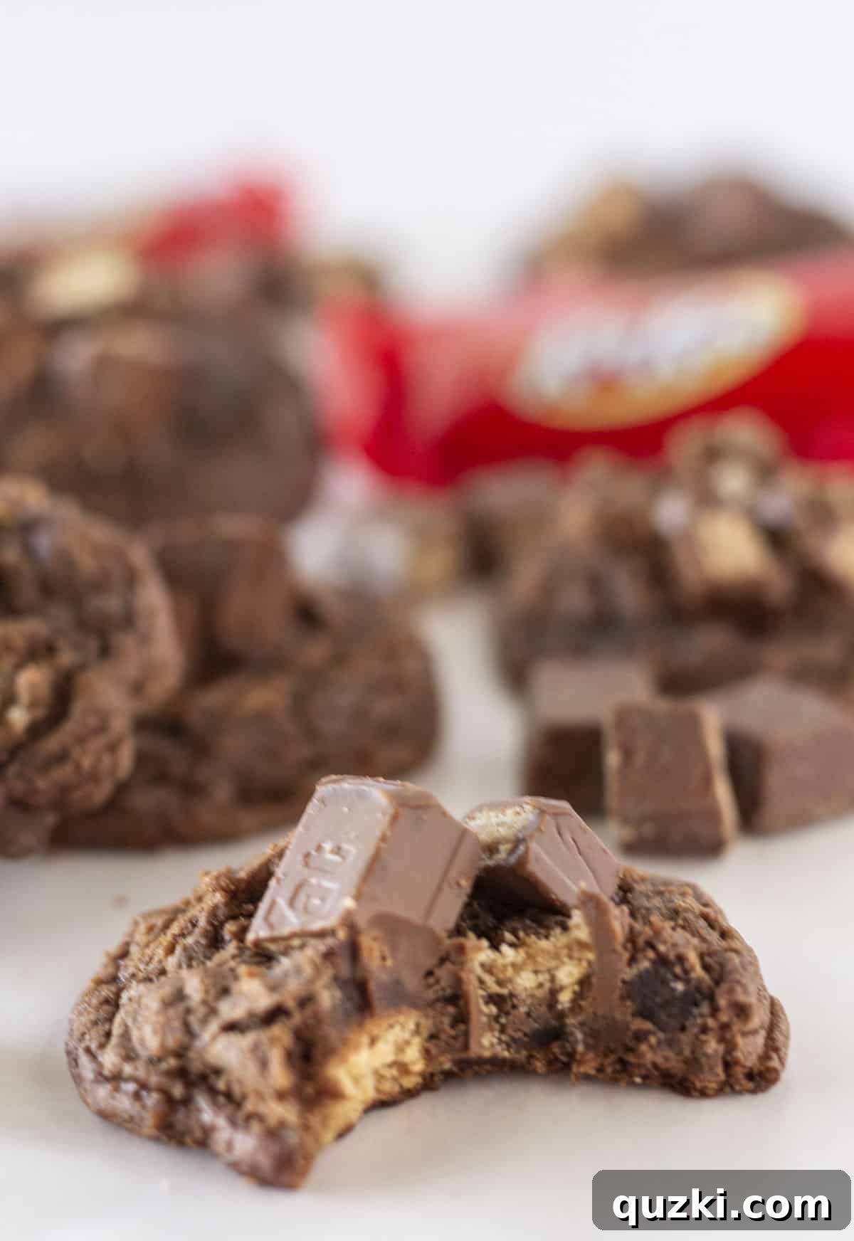 A delicious Kit Kat brownie cookie with a bite taken out, showing its chewy interior, with more cookies blurred in the background.