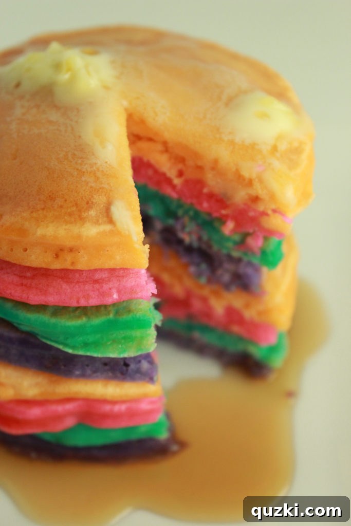 Top-down view of rainbow pancakes stacked on a plate with butter and syrup, surrounded by a festive table setting