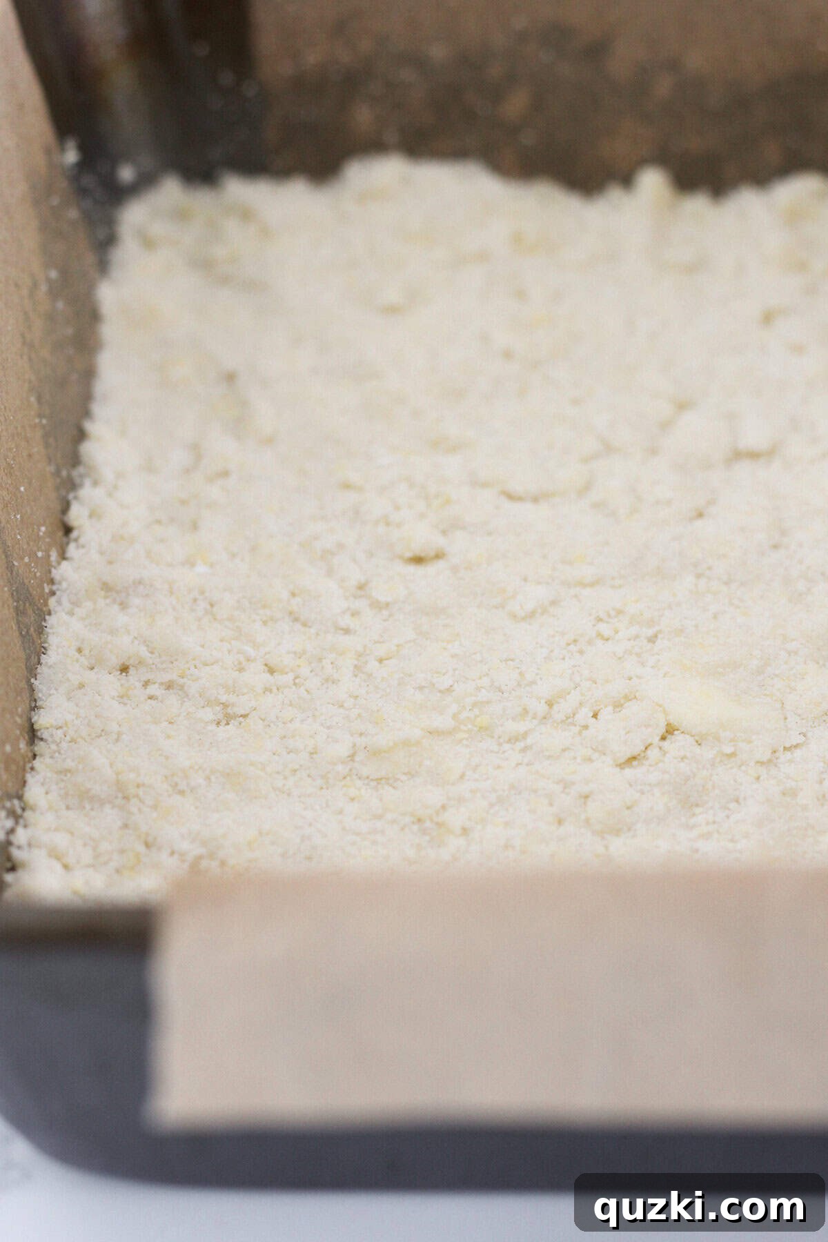 Half of the crumbly shortbread mixture being pressed evenly into a parchment-lined baking pan with fingertips, forming the base of the crumble bars.