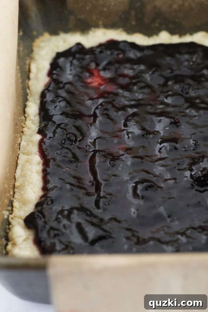 Blackberry jam being spread evenly over the warm, partially baked shortbread crust, leaving a small border around the edges.