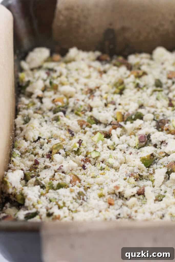 The remaining shortbread mixture, now combined with chopped pistachios, being sprinkled over the blackberry jam layer, forming the crumble topping.