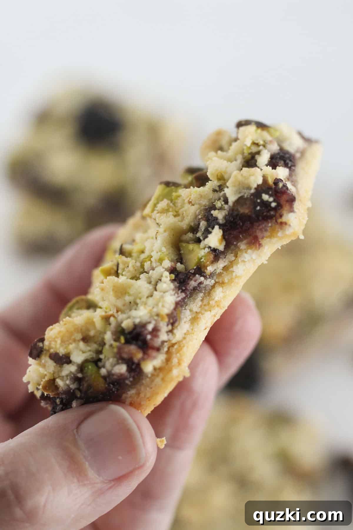 A single Blackberry Crumble bar with pistachios being held, showing a bite removed to reveal the luscious blackberry jam filling and crumbly layers.