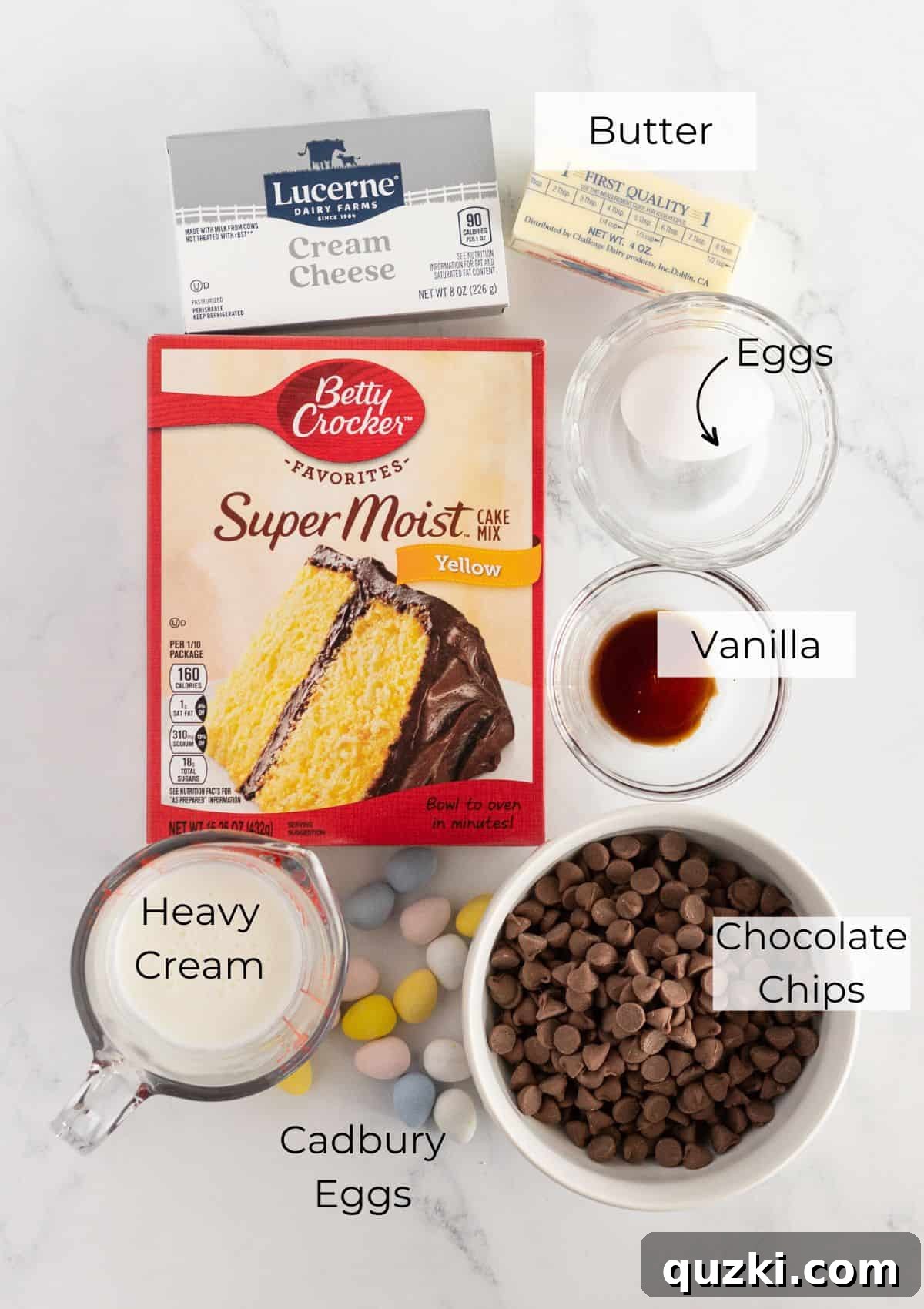 Quick-Bake Cadbury Mini Egg Cookies 3 A flat lay photograph displaying all the essential ingredients for baking Cadbury Mini Egg Cookies: a box of yellow cake mix, softened cream cheese and butter, a single egg, vanilla extract, milk chocolate chips, heavy cream, and a generous bag of Mini Cadbury Eggs.