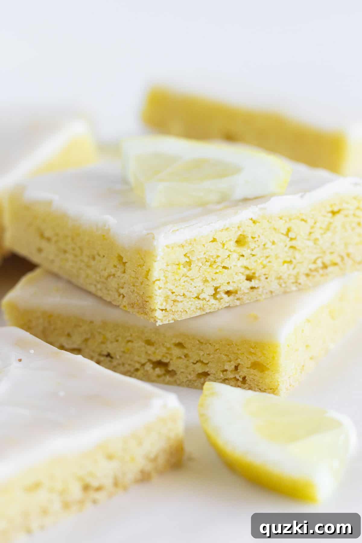A stack of golden, glazed lemon cake mix cookie bars on a white serving tray, garnished with fresh lemon zest.