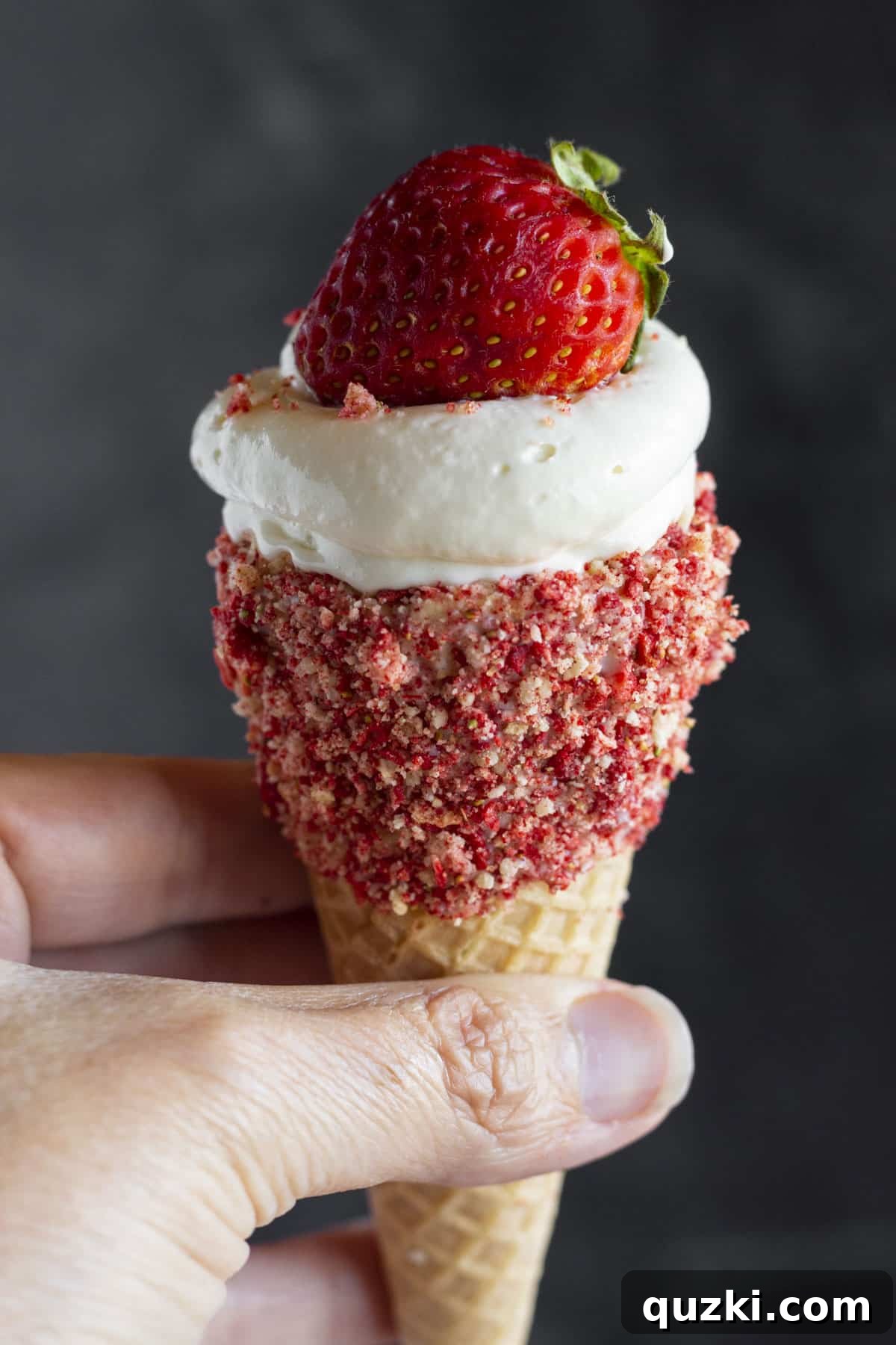 A hand holding a beautifully decorated Strawberry Crunch Cheesecake Cone, showcasing its tempting layers.