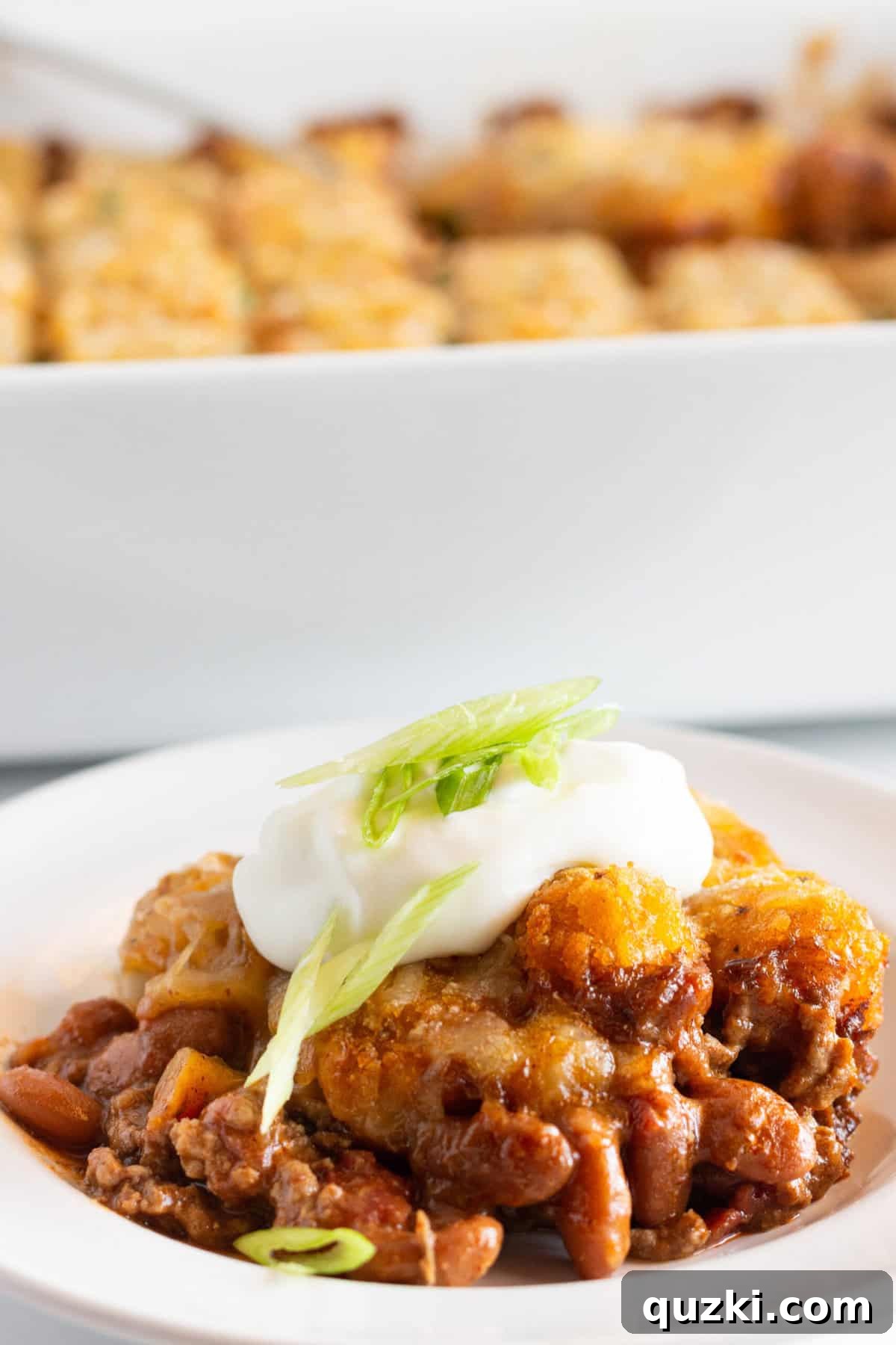 Hearty Chili Tater Tot Bake 5 A serving of Chili Cheese Tater Tot Casserole with sour cream and green onion toppings and the casserole in the background.