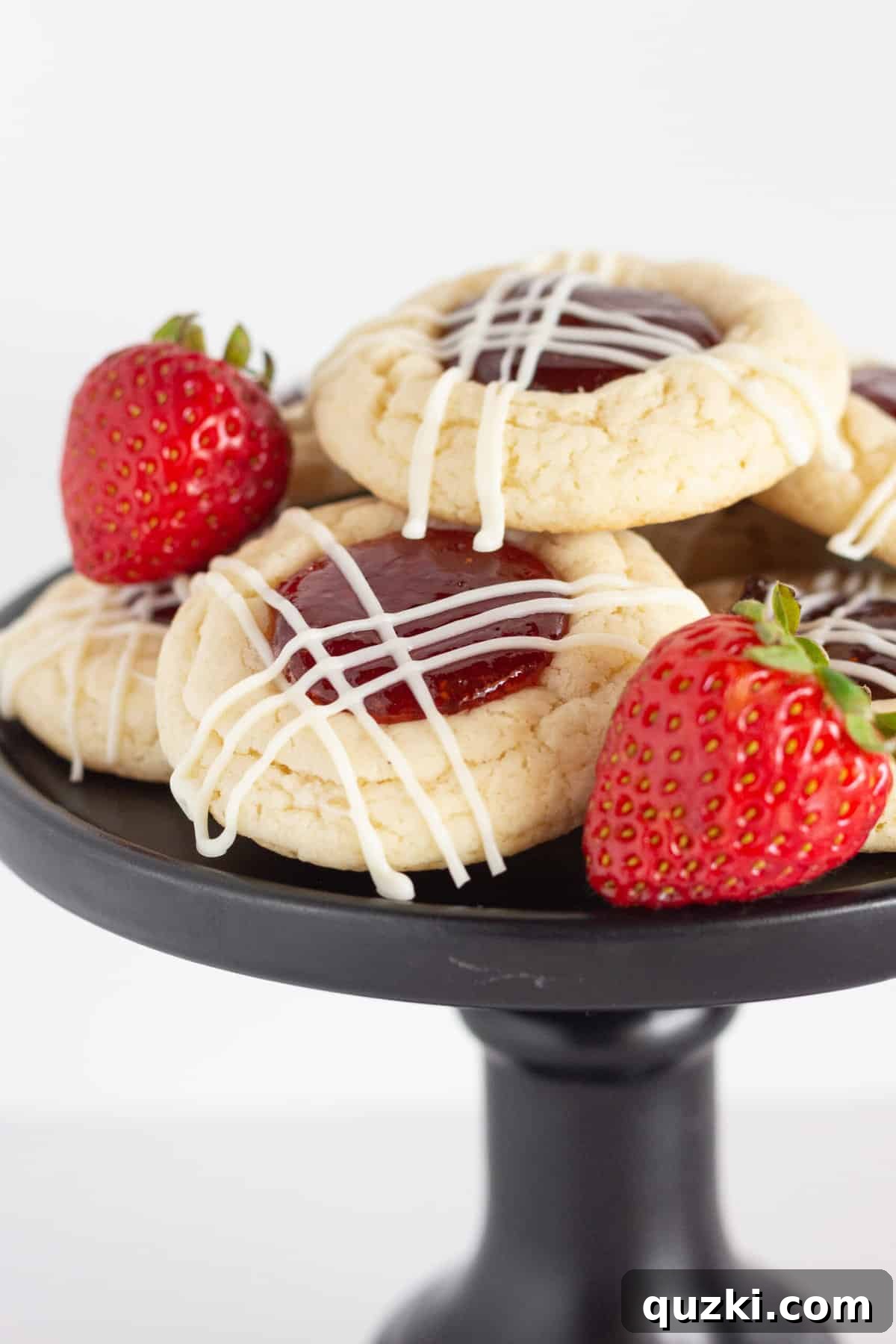 7 Effortless and Amazing Thumbprint Cookies 6 Easy Strawberry Jam Cookies artfully arranged on a black cake plate, each cookie beautifully drizzled with white chocolate, with a couple of fresh, ripe strawberries accompanying them.
