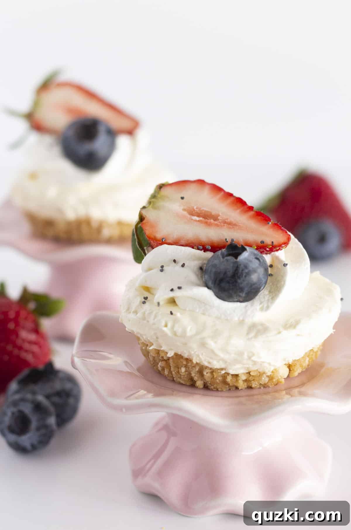 Quick No-Bake Cheesecake Bites 8 Two perfectly portioned mini no-bake cheesecakes, elegantly presented on small pink cake stands, garnished with an assortment of vibrant fresh berries.