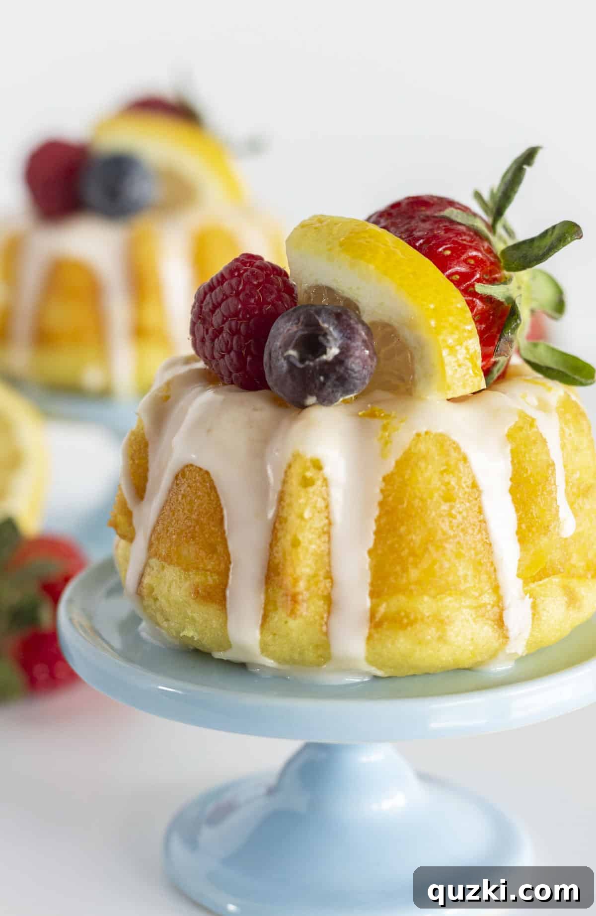 Two individual mini lemon bundt cakes, beautifully glazed and adorned with fresh berries, presented on charming blue mini plates.