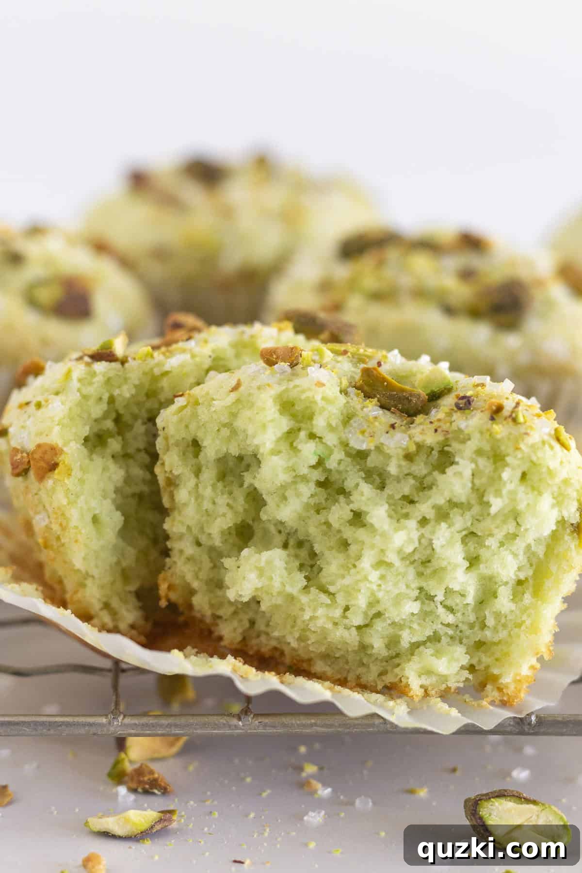 Pudding Mix Perfection: Effortless Pistachio Muffins 7 A pistachio muffin, removed from its paper liner and cut in half, revealing its tender green crumb, on a wire rack next to other whole muffins.