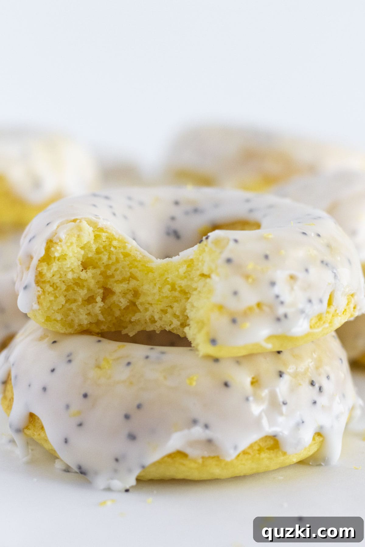 Oven-Baked Lemon Donuts with Zesty Poppy Seed Glaze 7 A close-up of a perfectly baked lemon donut with a bite taken, stacked enticingly atop another glazed donut, showcasing its moist texture and vibrant glaze.