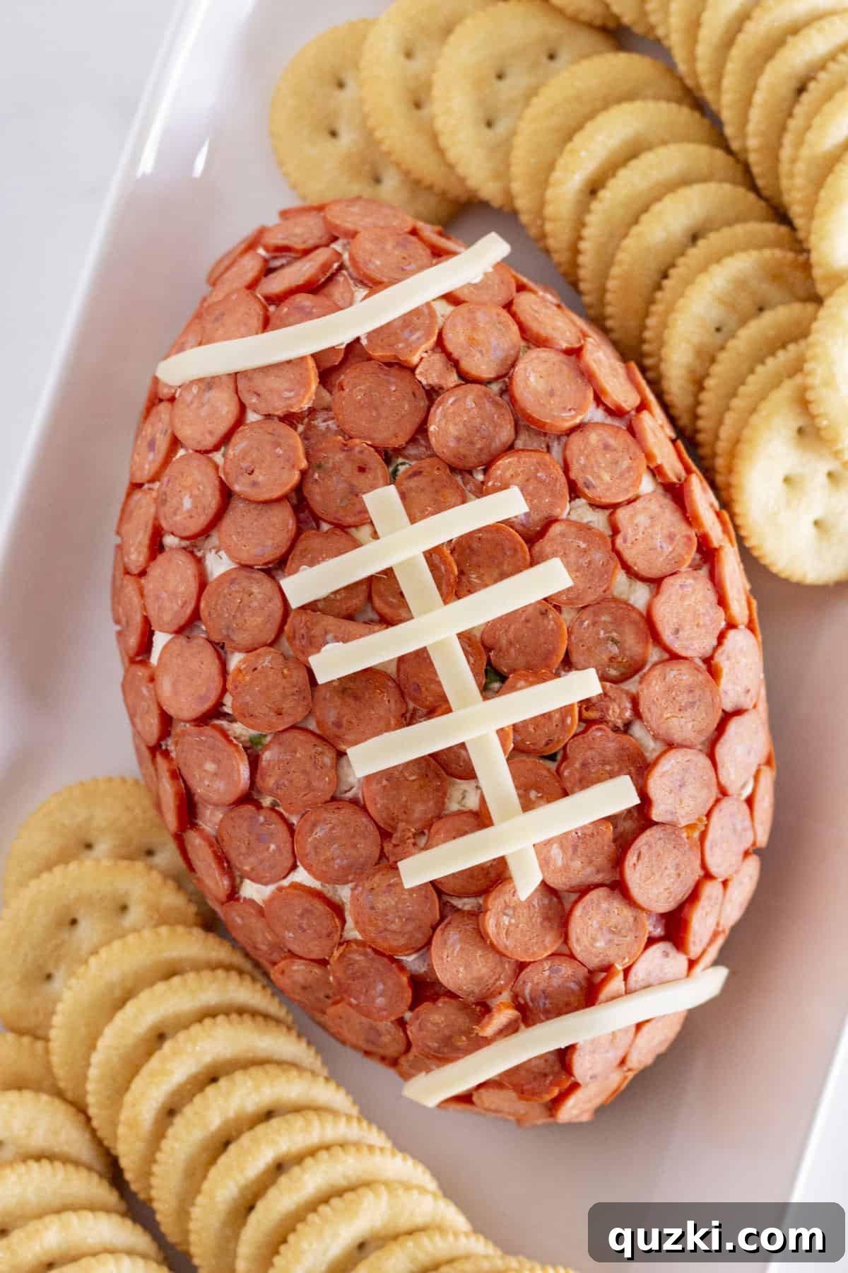 Touchdown Pizza Cheese Ball 2 A football-shaped cheese ball, meticulously decorated with mini pepperoni slices and distinctive string cheese laces, elegantly presented on a white platter alongside an assortment of crisp crackers for easy serving.
