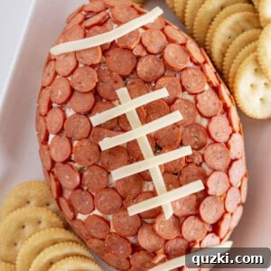 Touchdown Pizza Cheese Ball 8 A beautifully crafted football pepperoni cheese ball, perfectly shaped and adorned with cheese laces, rests on a pristine white serving tray surrounded by an array of crunchy crackers, ready to be enjoyed as a festive game day appetizer.