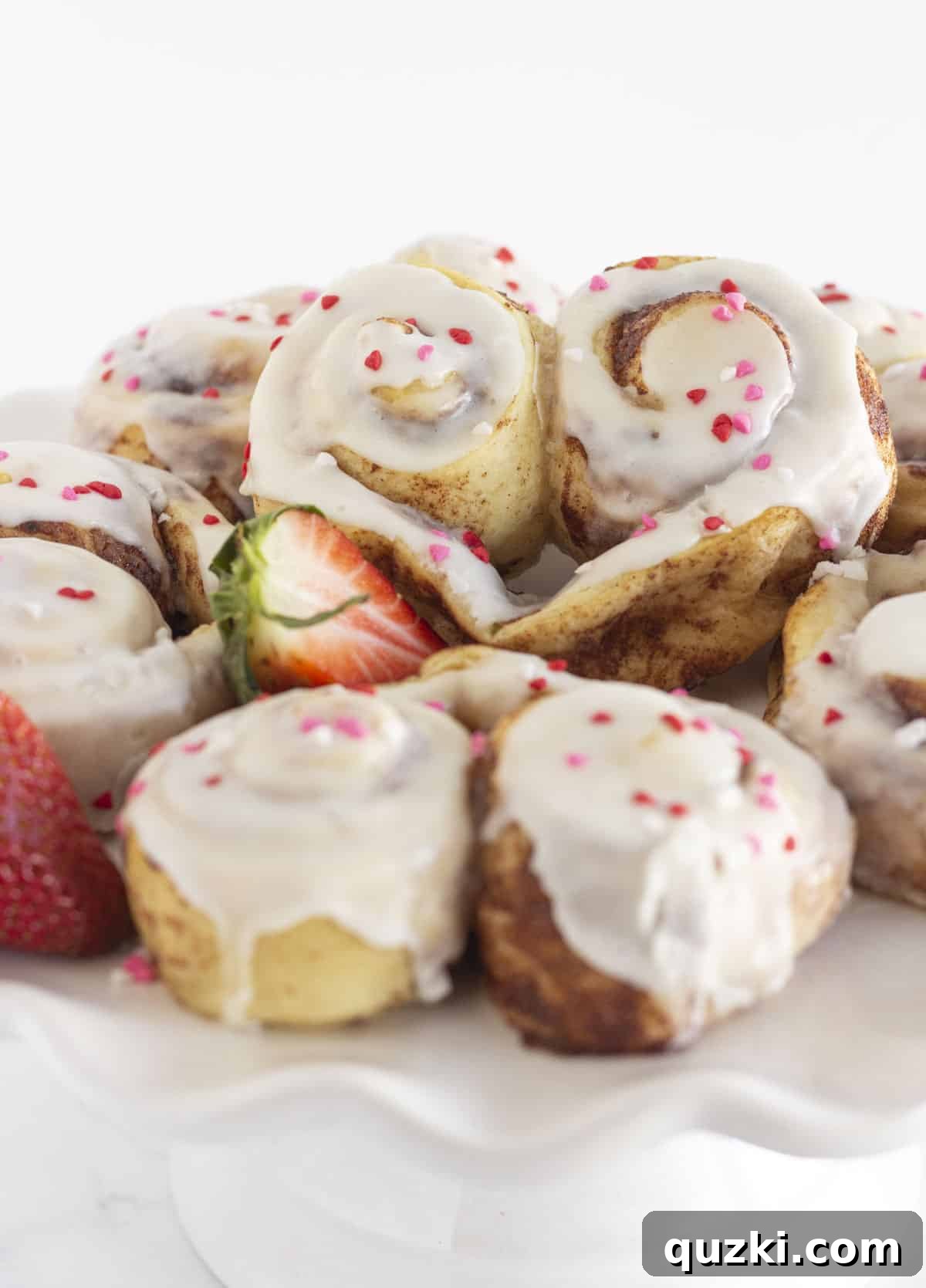 Simple & Sweet Heart Cinnamon Rolls 2 Heart shaped cinnamon rolls on a white cake plate, beautifully frosted and decorated with sprinkles.