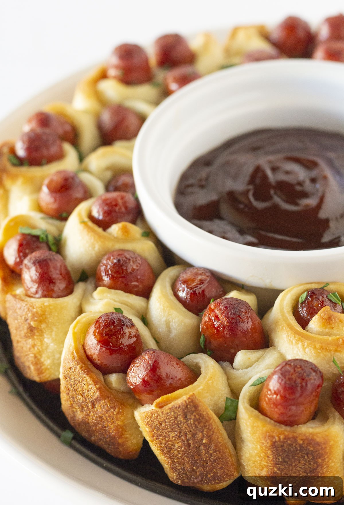 Golden-brown Pigs in a Blanket Wreath, baked to perfection with a small bowl of savory BBQ dipping sauce nestled elegantly in the center.