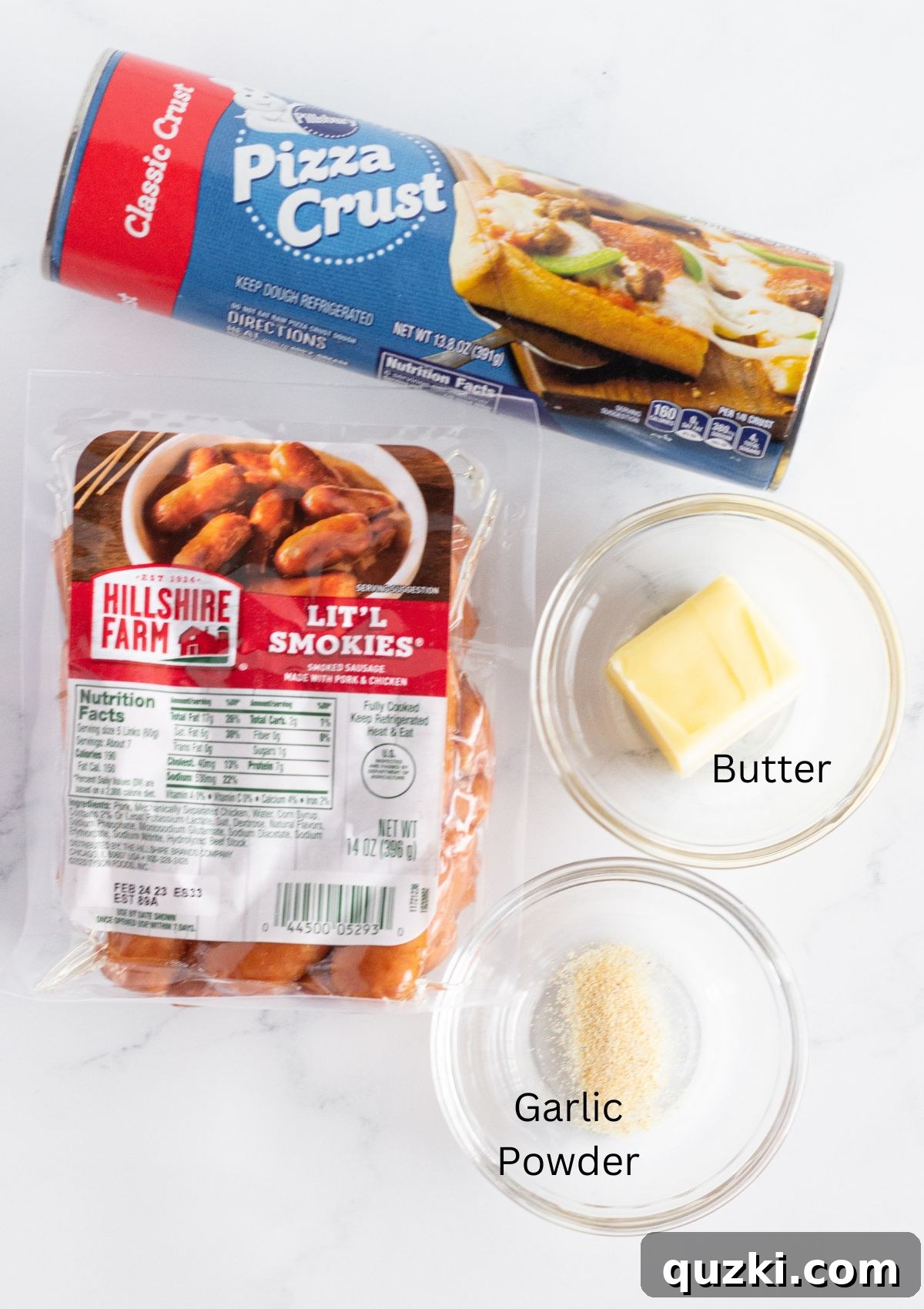 A well-arranged photograph displaying the four core ingredients required to prepare a wreath-shaped pigs in a blanket appetizer: refrigerated pizza dough, cocktail sausages, melted butter, and a container of garlic powder.