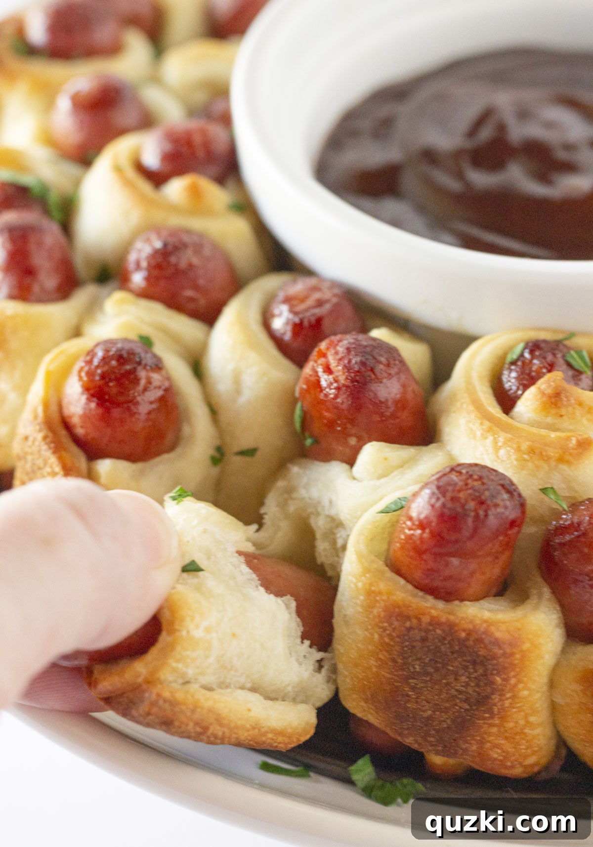 Close-up shot of a hand reaching in to pull a golden-brown pig in a blanket from the wreath, with a small bowl of BBQ dipping sauce in the center, ready for sharing at a party.