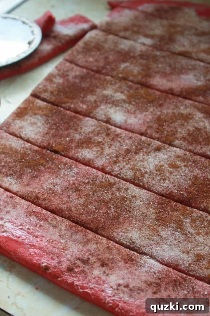 Red velvet dough rolled flat and evenly cut into long strips, adorned with a sweet cinnamon sugar filling, ready for assembly.