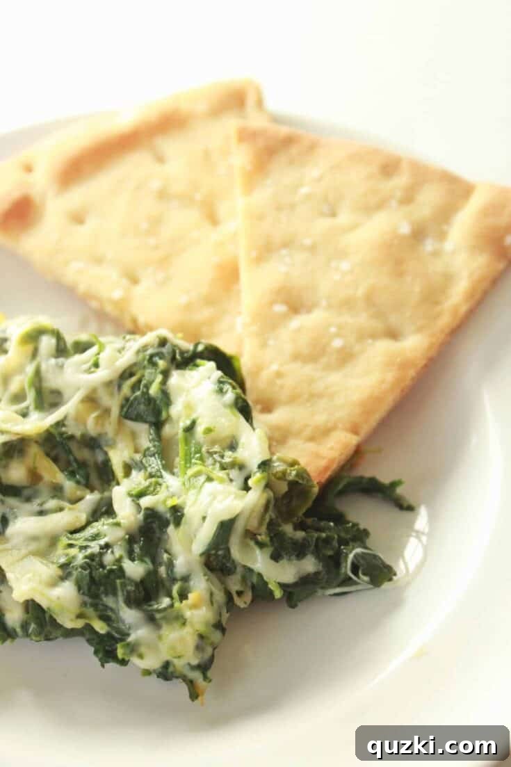 Warm and bubbly Spinach and Artichoke Dip, garnished with fresh herbs, served alongside a pile of perfectly crisp and seasoned homemade pita chips for dipping.