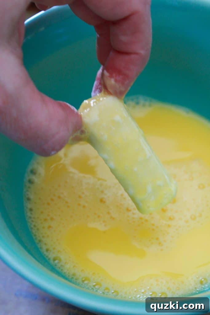 A floured mozzarella stick being dipped into a bowl of whisked egg wash.