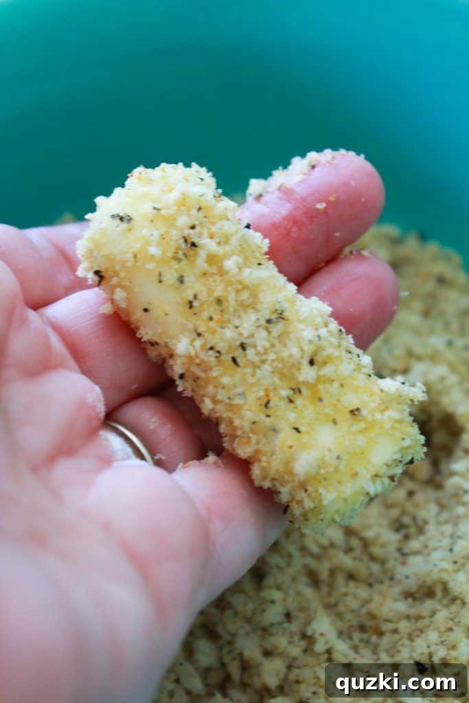 An egg-coated mozzarella stick being rolled and pressed into a bowl of seasoned panko breadcrumbs.
