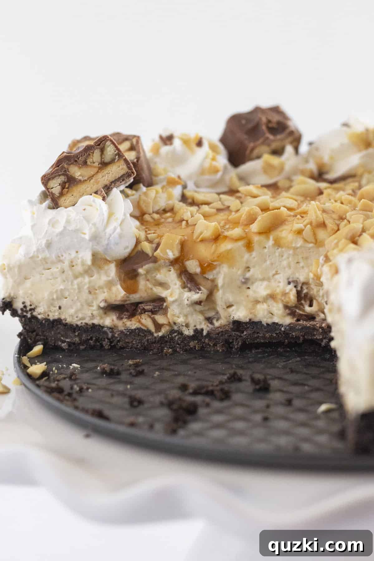 A close-up view of a beautifully prepared Snickers no-bake cheesecake, with a few slices already removed, revealing its delicious layered interior of creamy filling, chopped Snickers, caramel, and an Oreo crust.