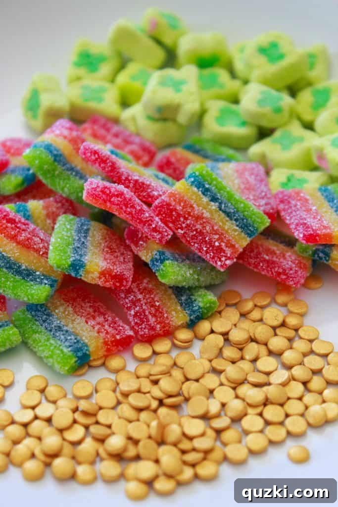 Lucky Leprechaun No-Bake Bark 3 A flat lay image displaying the colorful ingredients needed for Leprechaun Bark, including white chocolate, rainbow candies, clover marshmallows, and gold sprinkles.