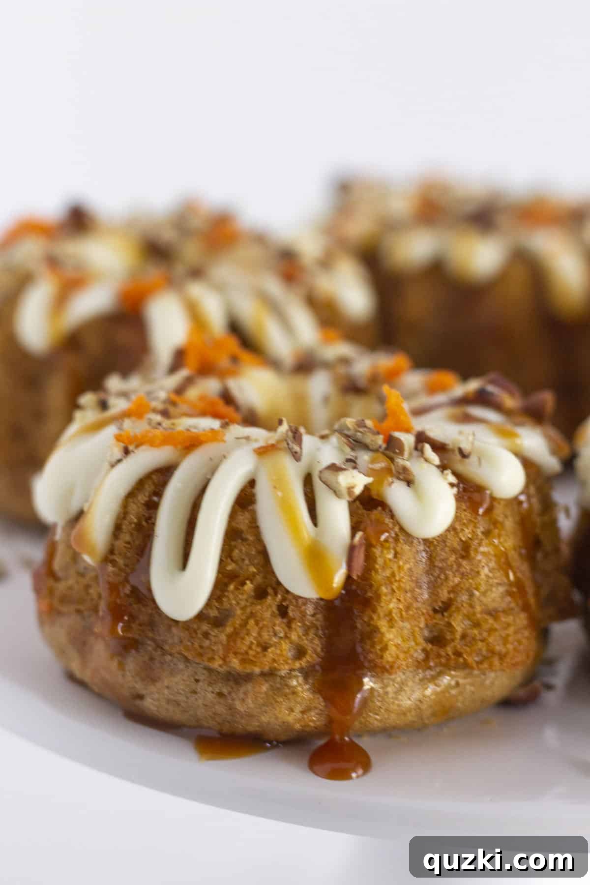 Effortless Mini Carrot Bundts 2 Mini carrot bundt cakes elegantly arranged on a white cake plate, adorned with frosting, caramel drizzle, and pecans.