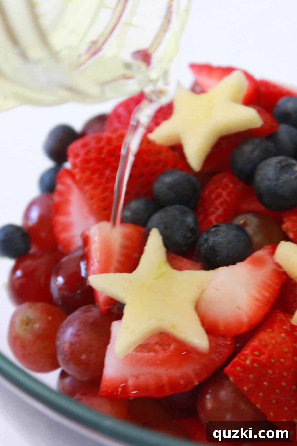 Patriotic Fruit Medley 6 A hand pouring translucent lime simple syrup over a bowl of mixed red, white, and blue fruits.