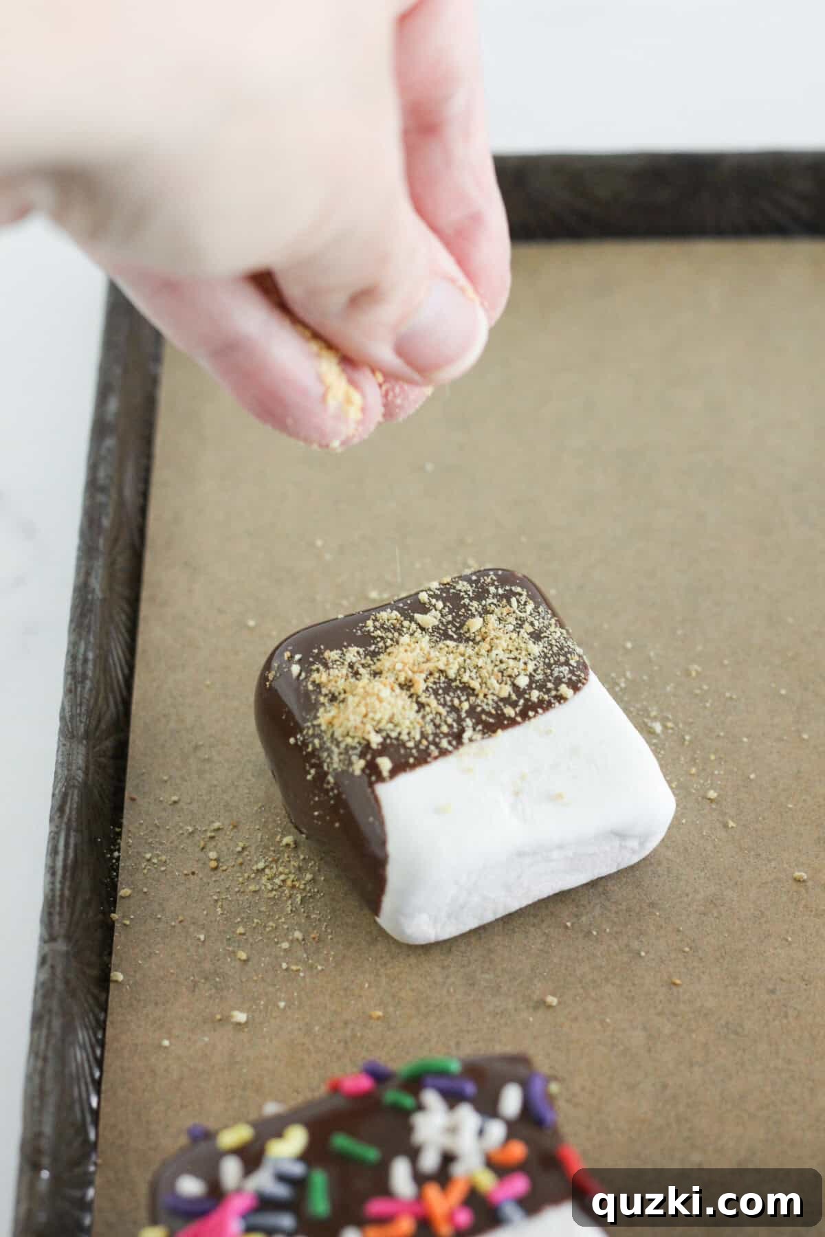 Easy Two-Ingredient Chocolate Dipped Marshmallows 6 A chocolate covered marshmallow being sprinkled with graham cracker crumbs, demonstrating the topping application process.
