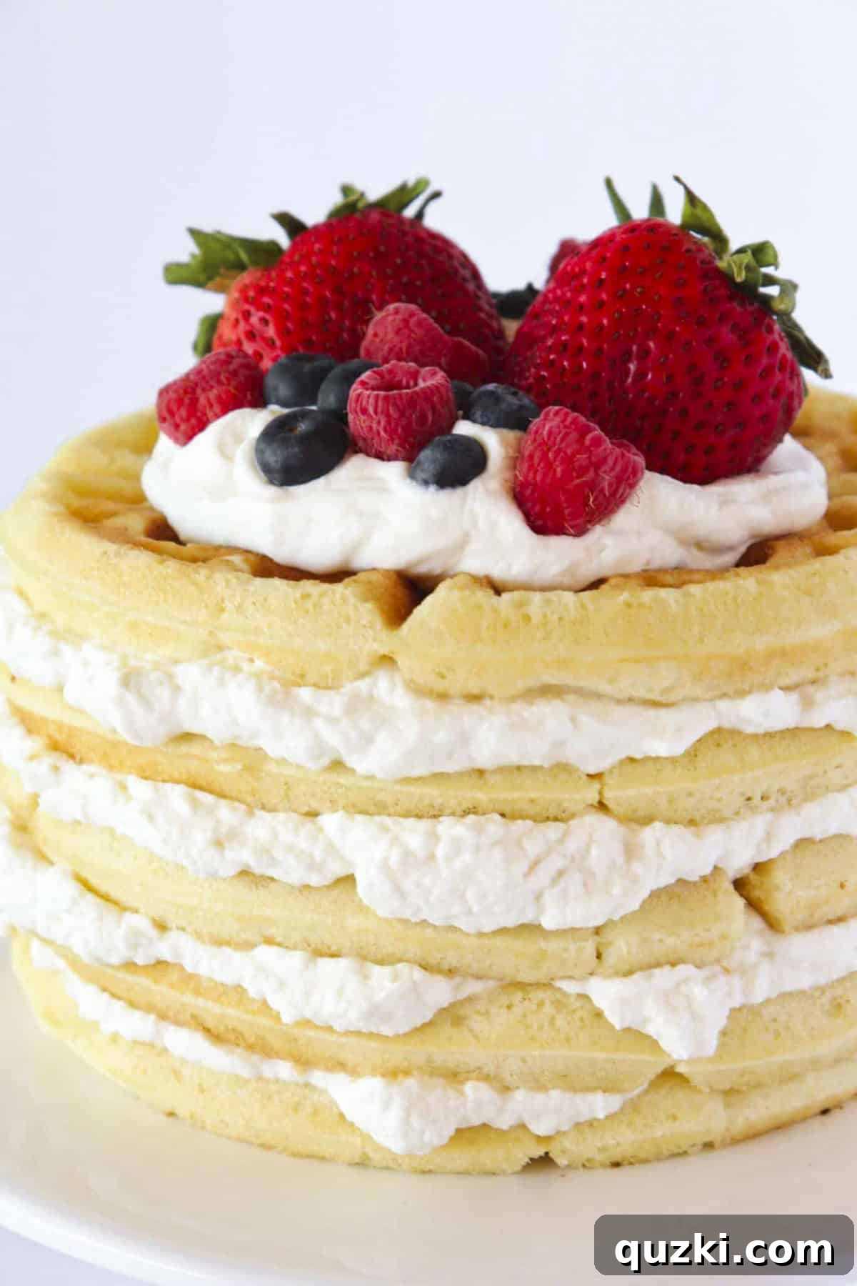 Towering Vanilla Waffle Cake 2 A beautifully layered vanilla waffle cake adorned with homemade whipped cream and an assortment of fresh strawberries, raspberries, and blueberries.