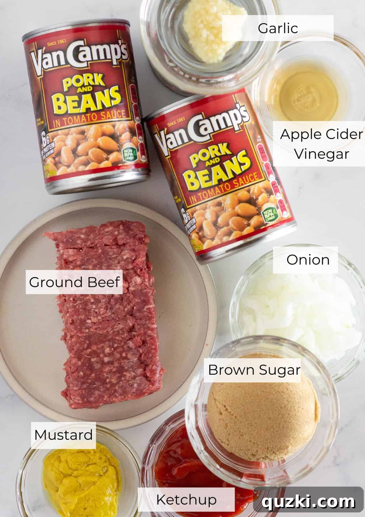 Hearty Ground Beef Baked Beans 3 A selection of fresh and pantry ingredients laid out on a wooden surface, including ground beef, a yellow onion, garlic cloves, cans of pork and beans, ketchup, brown sugar, apple cider vinegar, and yellow mustard, ready for making baked beans with hamburger.