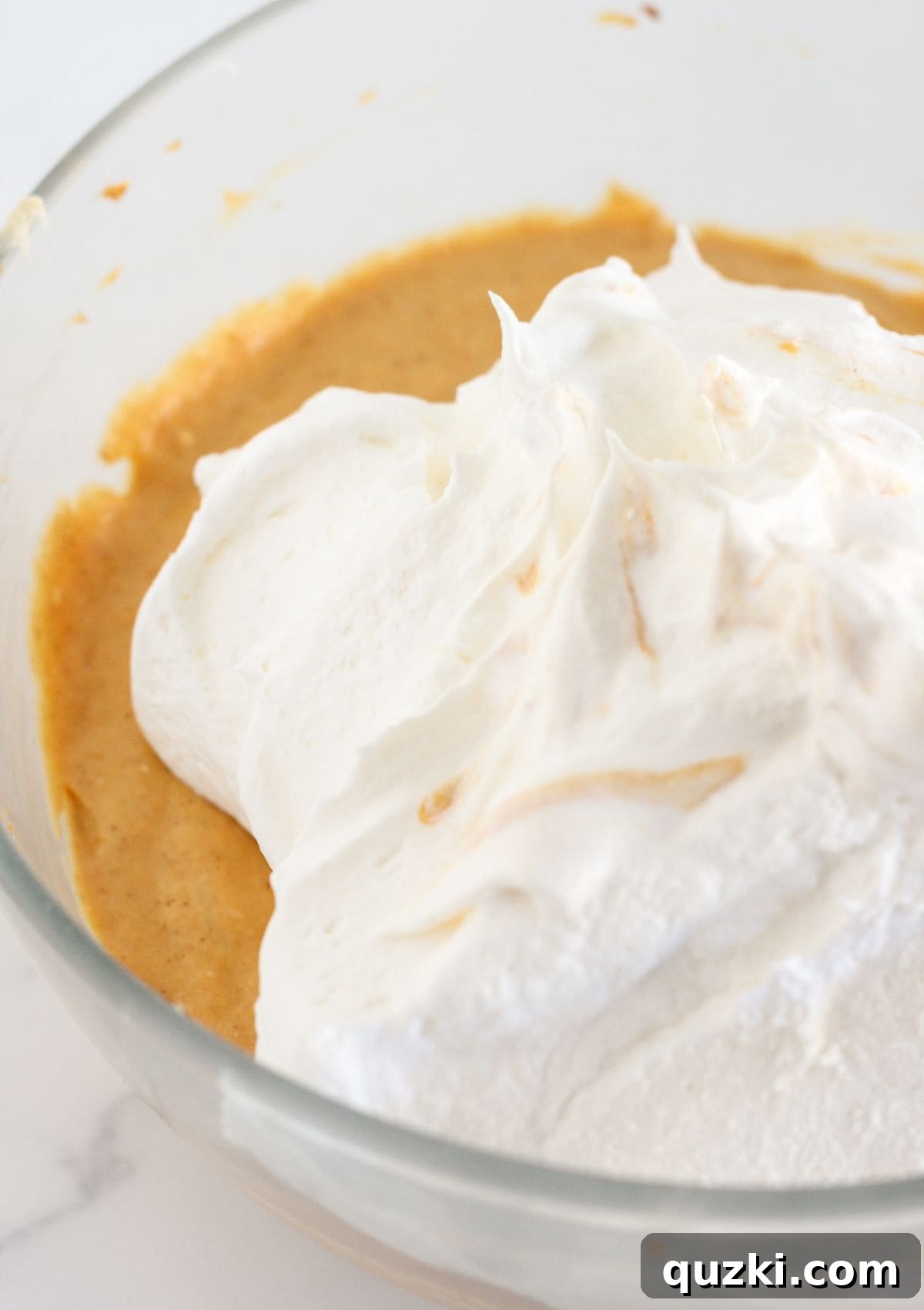 The fluffy whipped topping is gently being folded into the pumpkin mixture with a spatula, creating the light and airy mousse.