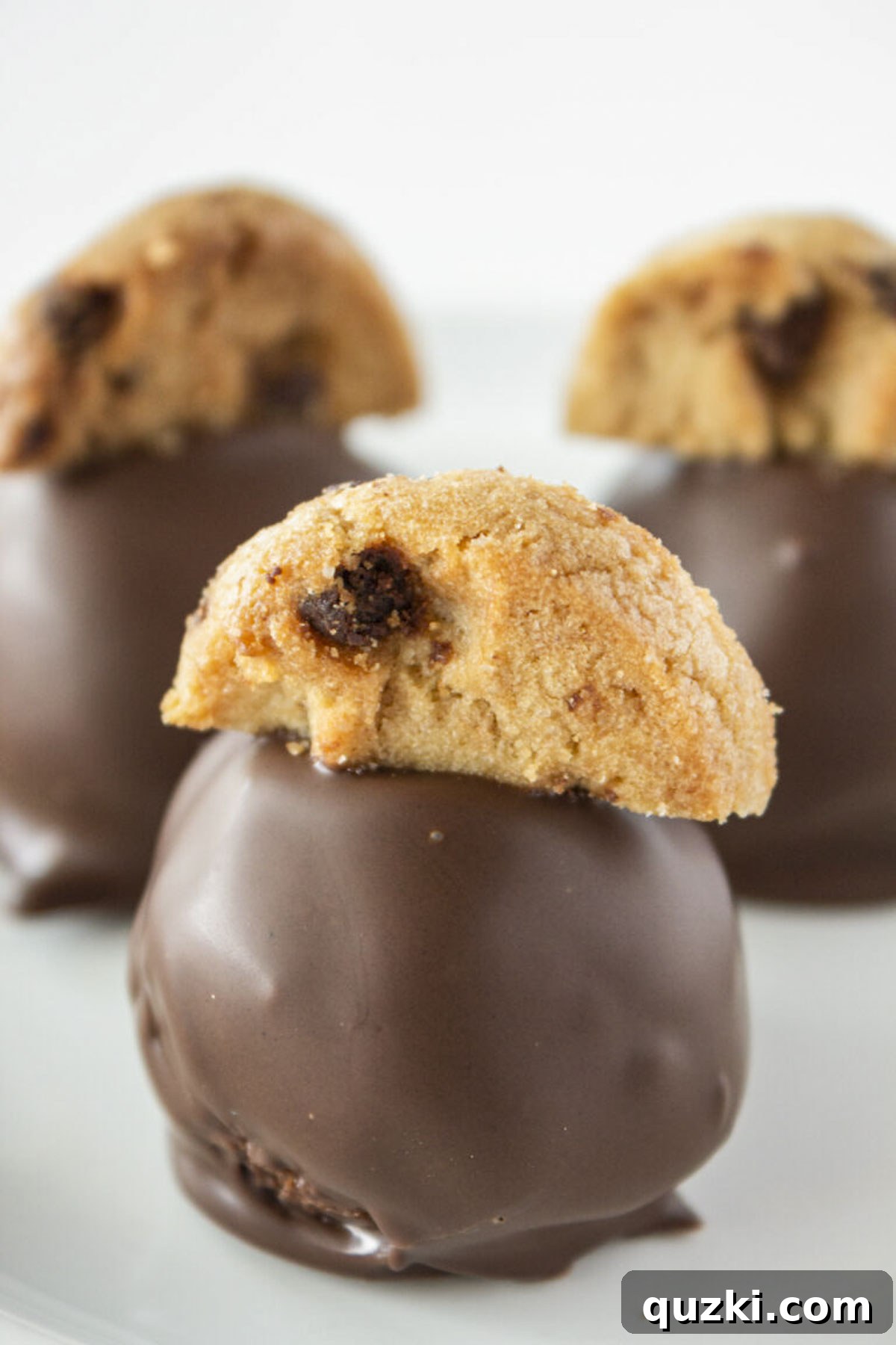 Three chocolate chip cookie truffles on a white plate, showcasing their rich chocolate coating and cookie garnish.
