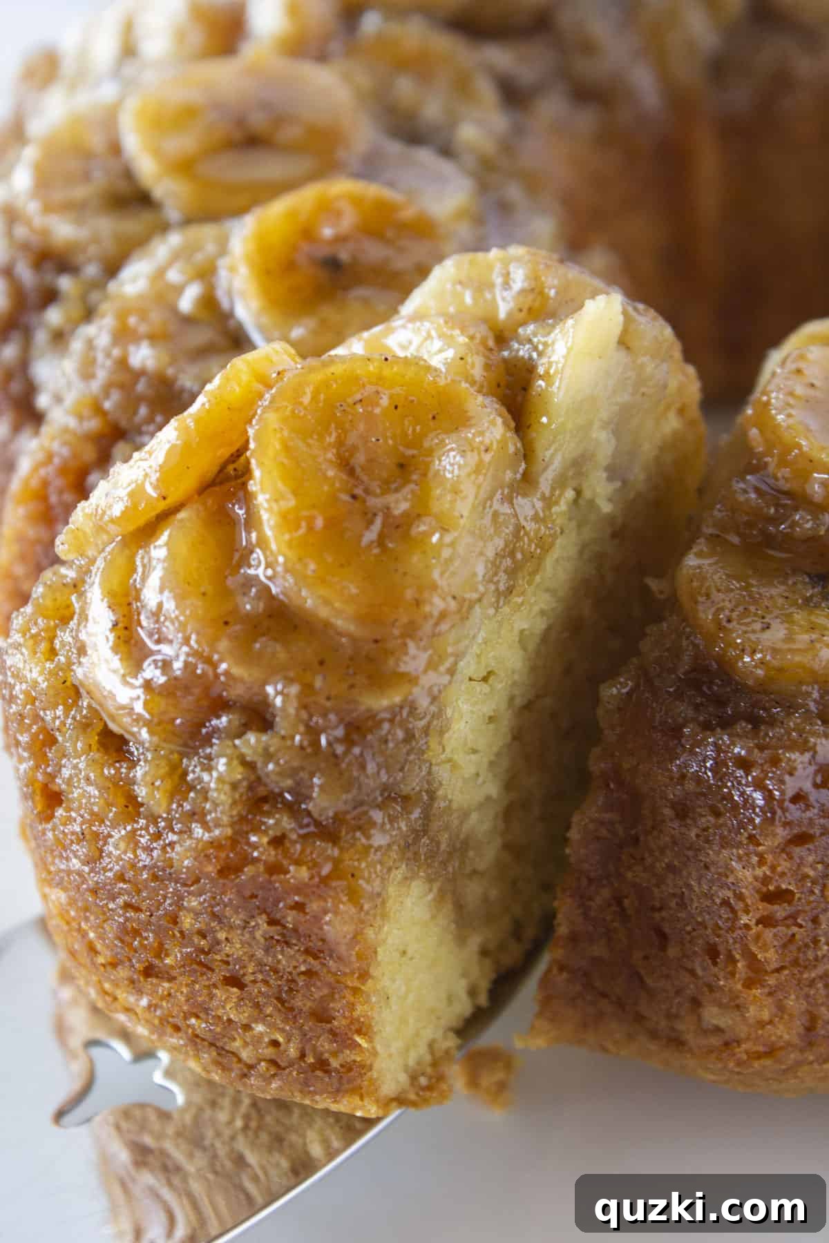 Moist Banana Bundt Magic 2 A close-up photo of a hand serving a perfect slice of warm banana upside down cake from a cake stand.