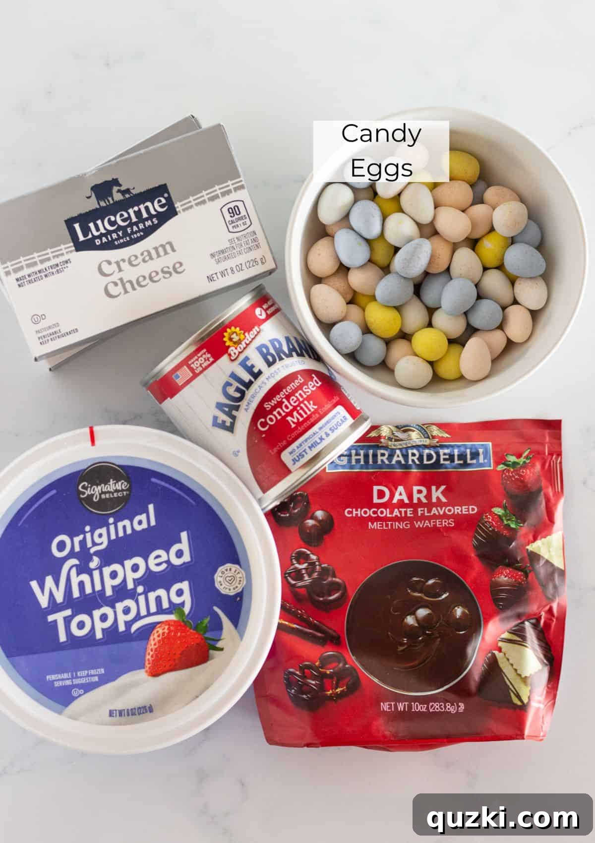All the necessary ingredients laid out for preparing the Mini Egg No-Bake Cheesecake, including cream cheese, sweetened condensed milk, whipped topping, chocolate chips, and Cadbury Mini Eggs.