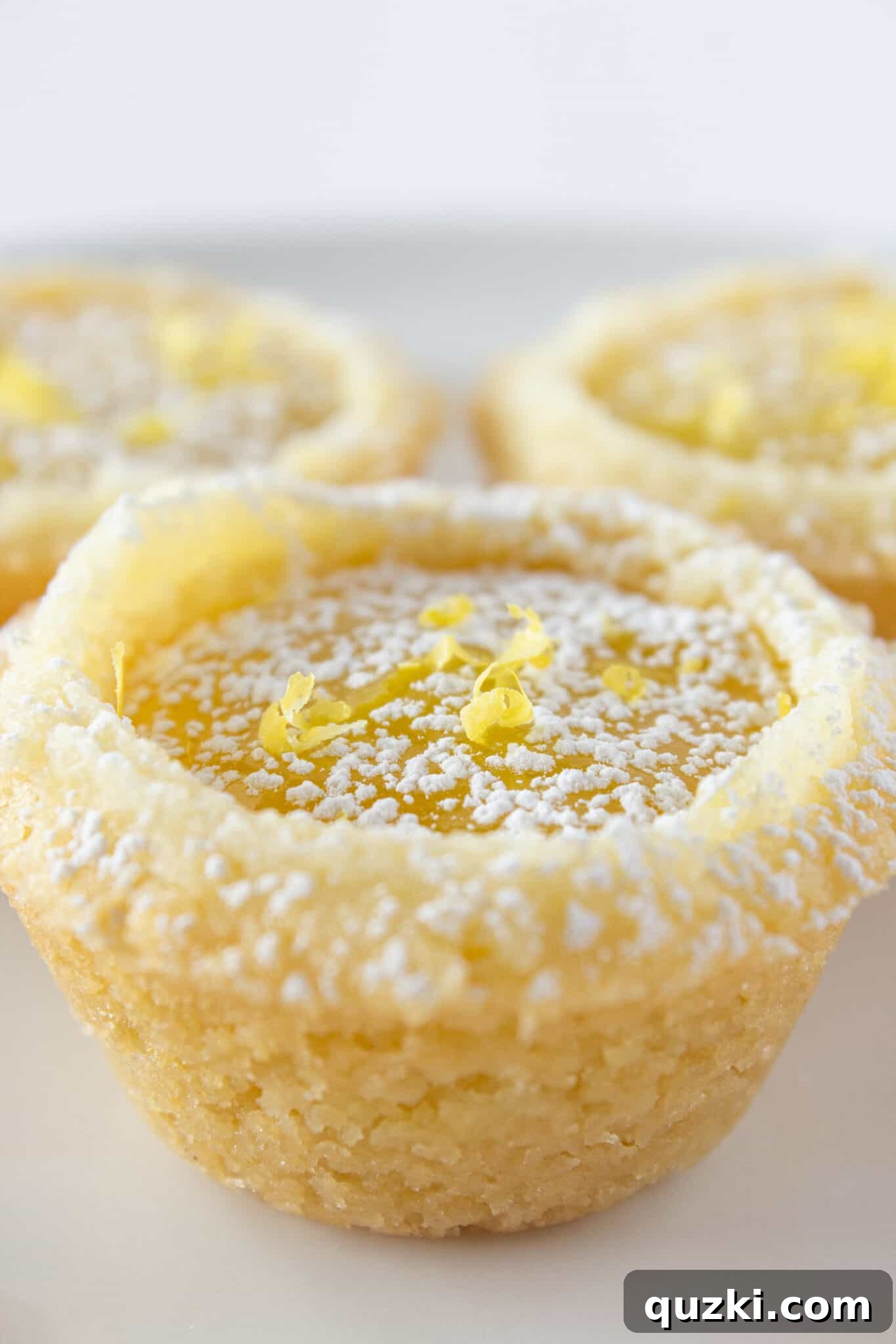 Three beautifully presented Lemon Bar Cookie Cups on a pristine white plate, garnished with a dusting of powdered sugar and a touch of fresh lemon zest.